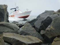 A rescue boat from a fire department is navigating through a rocky sea area with visible splashes of water. It is surrounded by large boulders along a coastline under a cloudy sky.