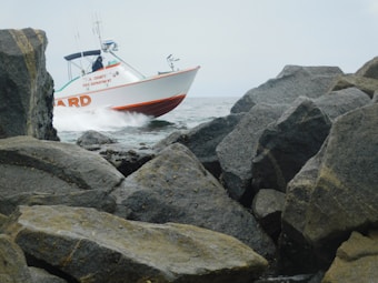 A rescue boat from a fire department is navigating through a rocky sea area with visible splashes of water. It is surrounded by large boulders along a coastline under a cloudy sky.