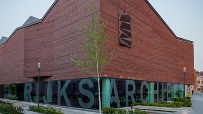 A large contemporary building with a distinct angular brick facade. The structure features large, bold letters on the ground floor glass windows, partially spelling out 'RIJKSARCHIEF.' A modern light fixture is installed on the wall, and a small tree is planted nearby. The sky is clear, and the building is surrounded by a well-maintained pedestrian area with greenery.