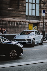 A silver luxury car is parked on a city street beside a large historic building with stone walls. Another black car is passing by in the foreground. Pedestrians are visible in the background near a street sign indicating parking and a poster.