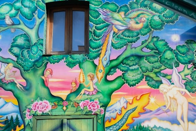 A vibrant mural features a whimsical scene with a mix of fantasy elements. A large tree with lush green leaves hosts a colorful parrot perching on its branches. Various mythical creatures, resembling fairies, are seated on the tree, surrounded by bright flowers. A dragon's tail curves along the bottom, above a landscape of mountains and a glowing sun or moon. The scene is framed by a window set into the wall.