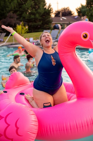 A person in a blue swimsuit is joyfully sitting on a large, inflatable pink flamingo float in a pool. The background shows several other people in the pool, some holding drinks, and a grassy area with lounge chairs. The atmosphere appears lively and festive.