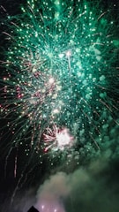 A vibrant display of fireworks lighting up the night sky with bright green and red colors, accompanied by smoke billowing at the bottom. The intricate patterns of the exploding fireworks create a dynamic and festive atmosphere.