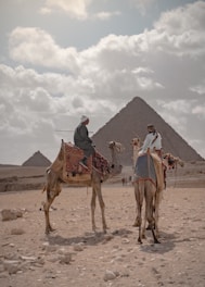 A desert scene with two camels carrying riders, set against the backdrop of pyramids on a cloudy day. The camels are adorned with patterned blankets and the riders wear traditional attire.