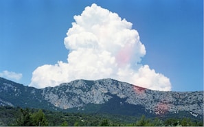 A large, fluffy white cloud rises dramatically above a rugged mountain range covered with greenery. The sky is bright blue, providing a clear contrast to the cloud and the textured gray and green mountains.