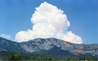 A large, fluffy white cloud rises dramatically above a rugged mountain range covered with greenery. The sky is bright blue, providing a clear contrast to the cloud and the textured gray and green mountains.