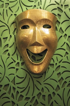 A gold theatrical mask with a smiling expression is mounted against a decorative green background featuring an intricate leaf-like pattern.