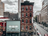 A cityscape featuring a prominent red brick building with a restaurant on the ground floor and a variety of other brick and modern buildings in the background. The street is lined with parked cars and trees, creating an urban environment with a slightly overcast sky.