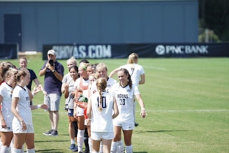 A group of female soccer players are gathered on a field, smiling and interacting with each other. They are wearing white jerseys with the word 'Hoyas' printed on them. A few people are in the background, including a man holding a camera and some other individuals who appear to be coaching staff or spectators.