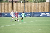 Three female soccer players are actively competing on a grassy field. One player in a burgundy uniform is in possession of the ball, trying to maneuver past a player in a bright green goalkeeper's uniform, while a player in a white uniform is approaching from the side. The scene is set against a backdrop of advertising banners.