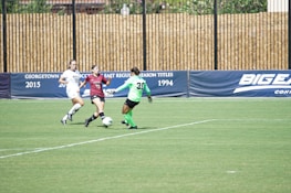 Three female soccer players are actively competing on a grassy field. One player in a burgundy uniform is in possession of the ball, trying to maneuver past a player in a bright green goalkeeper's uniform, while a player in a white uniform is approaching from the side. The scene is set against a backdrop of advertising banners.