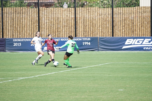 Three female soccer players are actively competing on a grassy field. One player in a burgundy uniform is in possession of the ball, trying to maneuver past a player in a bright green goalkeeper's uniform, while a player in a white uniform is approaching from the side. The scene is set against a backdrop of advertising banners.