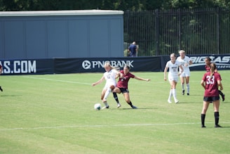A women’s soccer match in action on a grassy field with players wearing maroon and white jerseys. Two players are engaged closely, one trying to tackle the ball from the other. Other players are visible in the background, watching the play unfold. The scene is set against the backdrop of a fence and a large advertisement banner.