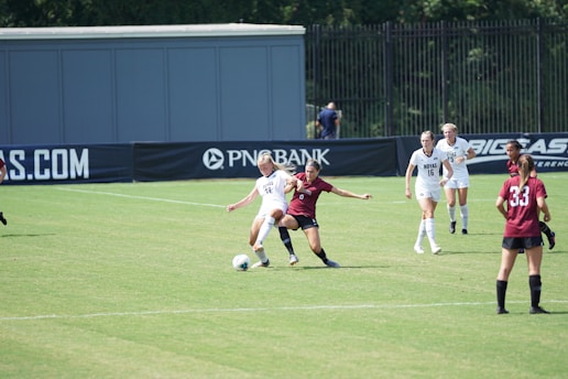 A women’s soccer match in action on a grassy field with players wearing maroon and white jerseys. Two players are engaged closely, one trying to tackle the ball from the other. Other players are visible in the background, watching the play unfold. The scene is set against the backdrop of a fence and a large advertisement banner.
