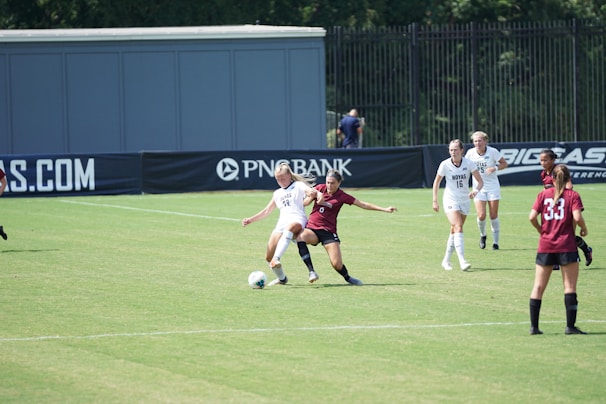 A women’s soccer match in action on a grassy field with players wearing maroon and white jerseys. Two players are engaged closely, one trying to tackle the ball from the other. Other players are visible in the background, watching the play unfold. The scene is set against the backdrop of a fence and a large advertisement banner.