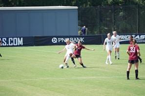 A women’s soccer match in action on a grassy field with players wearing maroon and white jerseys. Two players are engaged closely, one trying to tackle the ball from the other. Other players are visible in the background, watching the play unfold. The scene is set against the backdrop of a fence and a large advertisement banner.