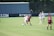 A women’s soccer match in action on a grassy field with players wearing maroon and white jerseys. Two players are engaged closely, one trying to tackle the ball from the other. Other players are visible in the background, watching the play unfold. The scene is set against the backdrop of a fence and a large advertisement banner.