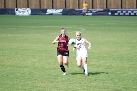 Two female soccer players are sprinting across a large green field, both focused on the soccer ball between them. One is wearing a maroon uniform with the number 22, and the other is in a white uniform with the number 18. In the background, a banner on the fence indicates a sporting event, and a person can be seen standing near the fence.