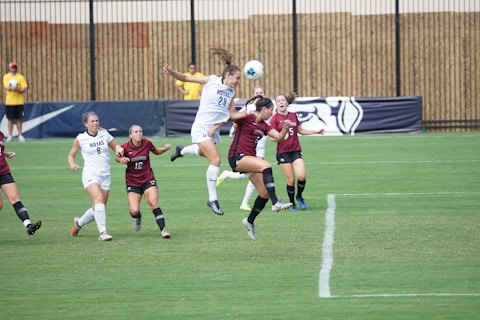 Young female soccer players are actively engaged in a competitive match on a grass field. One player in a white jersey is mid-air attempting to head the ball, while players in maroon jerseys and white jerseys surround her. A coach and other players can be seen in the background near the fence.