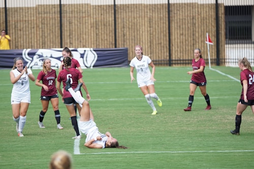 A group of women are playing a soccer game on a grassy field. Several players in maroon jerseys and some in white are actively engaged in the match. One player in white is performing a backflip or tumbling move while others are running and observing. A fence and a bulldog banner are visible in the background.
