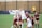 A women's soccer match is taking place with players actively competing for the ball in the air. One player, wearing a white uniform with the number 10, is leaping to head the ball. Surrounding her are several players in maroon uniforms, also attempting to gain control of the ball. The setting is an outdoor grass field with a wooden fence and banners in the background. A referee in a yellow shirt is observing the action from a distance.