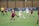 A women's soccer match is taking place on a grassy field, with players actively competing for control of the ball. Two teams are present, one in maroon jerseys and the other in white jerseys. A player in a white jersey is kicking the ball while surrounded by opponents. A referee in a yellow shirt is overseeing the game. A banner in the background highlights past achievements in soccer championships.