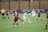 A women's soccer match is taking place on a grassy field, with players actively competing for control of the ball. Two teams are present, one in maroon jerseys and the other in white jerseys. A player in a white jersey is kicking the ball while surrounded by opponents. A referee in a yellow shirt is overseeing the game. A banner in the background highlights past achievements in soccer championships.