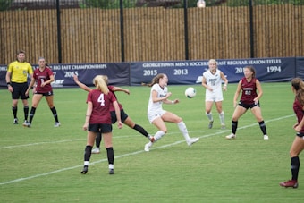 A women's soccer match is taking place on a grassy field, with players actively competing for control of the ball. Two teams are present, one in maroon jerseys and the other in white jerseys. A player in a white jersey is kicking the ball while surrounded by opponents. A referee in a yellow shirt is overseeing the game. A banner in the background highlights past achievements in soccer championships.