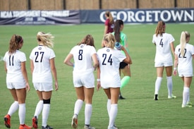 A group of female soccer players wearing white uniforms walk across a green field. Their backs are turned to the camera, and the players have numbers on their jerseys. A goalkeeper wearing a green uniform and pink socks is also visible. A banner in the background features the word 'Georgetown.'