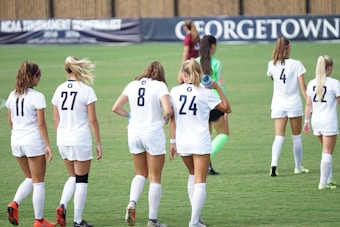 A group of female soccer players wearing white uniforms walk across a green field. Their backs are turned to the camera, and the players have numbers on their jerseys. A goalkeeper wearing a green uniform and pink socks is also visible. A banner in the background features the word 'Georgetown.'
