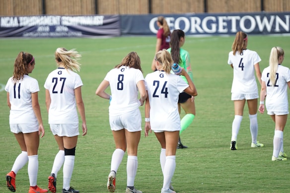 A group of female soccer players wearing white uniforms walk across a green field. Their backs are turned to the camera, and the players have numbers on their jerseys. A goalkeeper wearing a green uniform and pink socks is also visible. A banner in the background features the word 'Georgetown.'