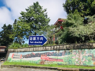 A sign in blue and white provides directional information to a park, situated in front of a lush, green environment. The background features a vivid mural depicting a red train and natural scenery. A traditional Asian structure with an ornate roof peeks through the dense greenery, contributing to the serene setting.