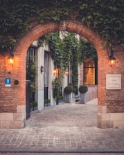 A charming and quaint cobblestone alleyway flanked by tall brick walls covered in vibrant green ivy. The entrance features an arched doorway with a carved stone face atop, and the soft glow of vintage-style lanterns illuminating the path. Potted plants line the walkway creating an inviting atmosphere. A sign indicates the presence of a hotel.