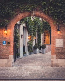 A charming and quaint cobblestone alleyway flanked by tall brick walls covered in vibrant green ivy. The entrance features an arched doorway with a carved stone face atop, and the soft glow of vintage-style lanterns illuminating the path. Potted plants line the walkway creating an inviting atmosphere. A sign indicates the presence of a hotel.