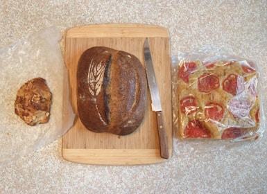 A wooden cutting board is placed on a speckled surface. On the board, there is a loaf of dark brown bread with a decorative flour pattern on top. Next to the bread is a serrated knife with a wooden handle. To the left of the board, there is a piece of nut-studded bread on wax paper. On the right, a packaged tomato-topped focaccia bread is visible, wrapped in plastic.