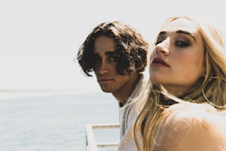 Two young adults, one with dark curly hair and the other with long blonde hair, are close together near a body of water. They both have serious expressions on their faces and are looking towards the camera. There is a blurred, bright background with a glimpse of the ocean under a clear sky.