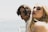 Two young adults, one with dark curly hair and the other with long blonde hair, are close together near a body of water. They both have serious expressions on their faces and are looking towards the camera. There is a blurred, bright background with a glimpse of the ocean under a clear sky.
