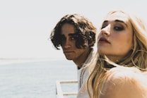 Two young adults, one with dark curly hair and the other with long blonde hair, are close together near a body of water. They both have serious expressions on their faces and are looking towards the camera. There is a blurred, bright background with a glimpse of the ocean under a clear sky.