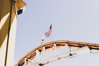 A section of a roller coaster track with an American flag and a California state flag flying at the top. The structure is primarily white with wooden supports and is captured against a clear sky. A part of a yellow structure is visible on the left side of the image.