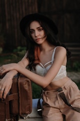 A woman with long dark hair and highlighted strands leans against a vintage suitcase. She is wearing a black hat, a white lace crop top, and plaid high-waisted pants with suspenders. The background is blurred, suggesting an outdoor setting, possibly near a wooden structure.