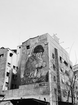 A large mural painted on the side of a building depicts a child sitting and looking down, with what appears to be a striped mask covering their eyes. The surrounding urban environment includes other buildings and leafless trees, creating a stark contrast with the detailed artwork.