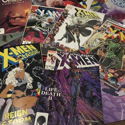 A collection of X-Men comic books spread out, featuring various covers and illustrations. Prominent characters like Storm are visible in dynamic poses with vibrant colors. The covers include titles like 'X-Men Classic' and 'The Uncanny X-Men' with distinct artwork styles and comic book logos.