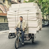 A man is riding a bicycle that is heavily loaded with stacked mattresses, navigating through a city street. He appears focused and determined, balancing the precarious load with care. Surrounding him are parked cars and trees lining the street, suggesting an urban environment.