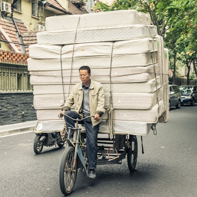 A man is riding a bicycle that is heavily loaded with stacked mattresses, navigating through a city street. He appears focused and determined, balancing the precarious load with care. Surrounding him are parked cars and trees lining the street, suggesting an urban environment.