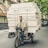 A man is riding a bicycle that is heavily loaded with stacked mattresses, navigating through a city street. He appears focused and determined, balancing the precarious load with care. Surrounding him are parked cars and trees lining the street, suggesting an urban environment.