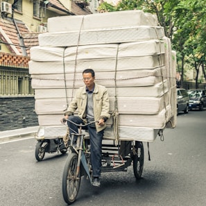 A man is riding a bicycle that is heavily loaded with stacked mattresses, navigating through a city street. He appears focused and determined, balancing the precarious load with care. Surrounding him are parked cars and trees lining the street, suggesting an urban environment.