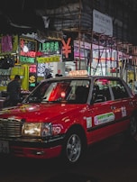 A red taxi is parked on a bustling city street lined with brightly lit neon signs. Various signs in multiple languages advertise currency exchange and sales. Scaffolding is visible on a nearby building, and a few pedestrians are walking.