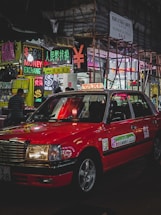 A red taxi is parked on a bustling city street lined with brightly lit neon signs. Various signs in multiple languages advertise currency exchange and sales. Scaffolding is visible on a nearby building, and a few pedestrians are walking.