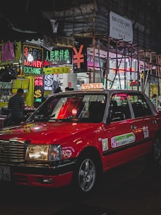 A red taxi is parked on a bustling city street lined with brightly lit neon signs. Various signs in multiple languages advertise currency exchange and sales. Scaffolding is visible on a nearby building, and a few pedestrians are walking.