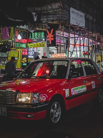 A red taxi is parked on a bustling city street lined with brightly lit neon signs. Various signs in multiple languages advertise currency exchange and sales. Scaffolding is visible on a nearby building, and a few pedestrians are walking.